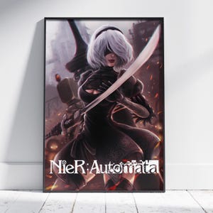 Peut inclure: Une affiche encadrée représentant un personnage aux cheveux blancs courts et au bandeau noir, portant une robe noire aux motifs complexes. Le personnage tient une longue épée en diagonale. Le texte NieR:Automata est imprimé en blanc en bas.