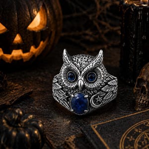 May include: A silver-toned ring shaped like an owl with detailed feather textures. The owl has two small, round, dark blue gemstone eyes and a larger, oval-shaped, dark blue gemstone set at the base, accompanied by a small silver crescent moon.