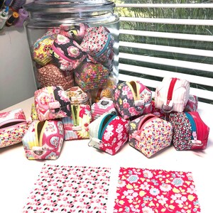 May include: A glass jar filled with colorful fabric coin purses. The purses are arranged in a pile on a white surface, with two additional fabric squares beside them. The fabric squares have a pink and white geometric pattern and a red and white floral pattern.