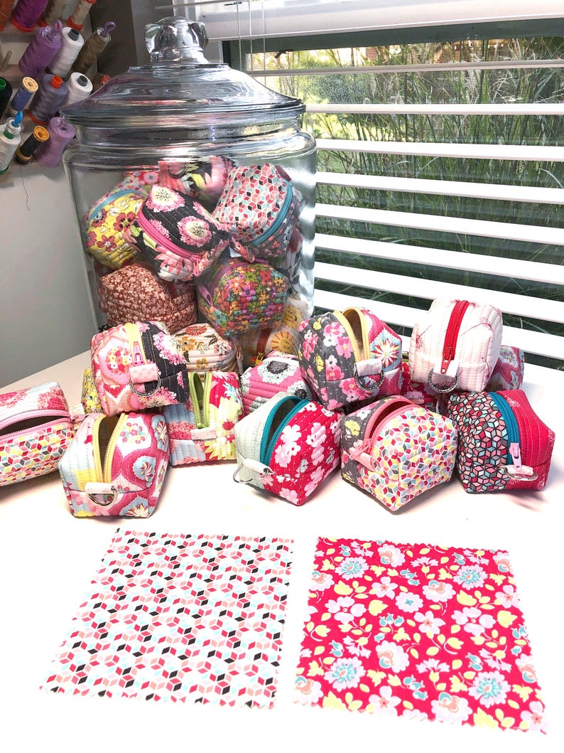 May include: A glass jar filled with colorful fabric coin purses. The purses are arranged in a pile on a white surface, with two additional fabric squares beside them. The fabric squares have a pink and white geometric pattern and a red and white floral pattern.