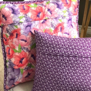 Puede incluir: Dos almohadas decorativas. Una almohada tiene un patr&oacute;n floral con flores rosas, moradas y verdes sobre un fondo blanco. La otra almohada es de color morado s&oacute;lido con un patr&oacute;n floral blanco.