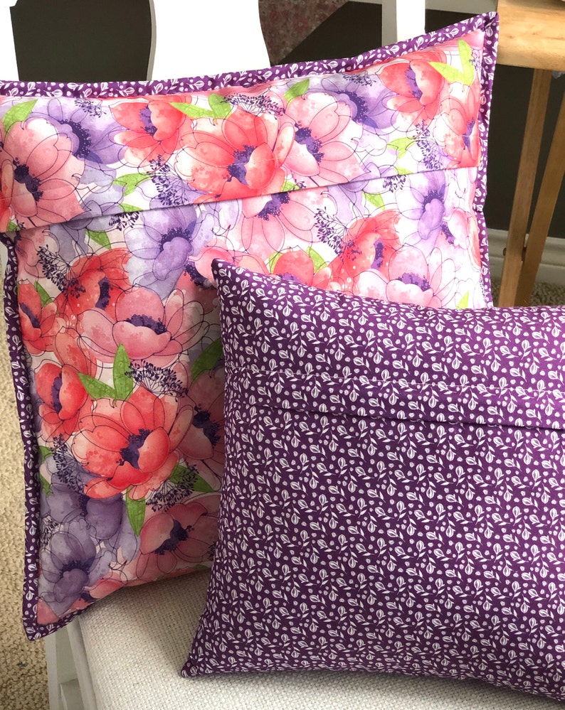 Puede incluir: Dos almohadas decorativas. Una almohada tiene un patr&oacute;n floral con flores rosas, moradas y verdes sobre un fondo blanco. La otra almohada es de color morado s&oacute;lido con un patr&oacute;n floral blanco.