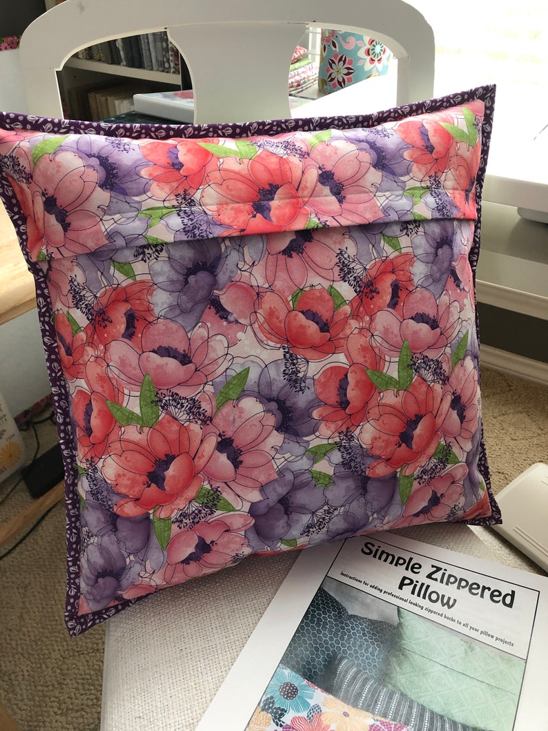 Puede incluir: Una almohada floral con un estampado floral morado y rosa. La almohada tiene una cremallera y est&aacute; hecha con un patr&oacute;n de almohada con cremallera simple. El texto en el patr&oacute;n dice "Simple Zippered Pillow. Instructions for adding professional looking zippered backs to all your pillow projects."