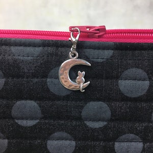 May include: A black zippered pouch with a silver moon and cat charm. The pouch has a pink zipper and a black and gray polka dot pattern.