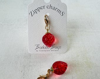 May include: Red strawberry zipper charms with gold-colored hardware. The charms are attached to a white tag with the text "Zipper charms" and the brand name "Bodobo Bags". The strawberry charms have a textured surface.