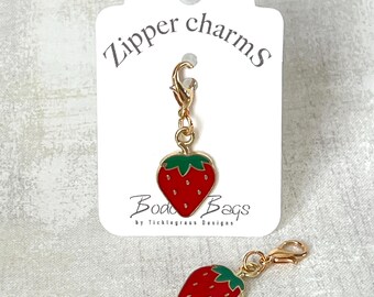 May include: Two gold zipper charms shaped like strawberries. The charms have red and green enamel and a gold lobster clasp. The charms are on a white card with the text "Zipper charms" and "Boo Boo Bags by Ticklegrass Designs".