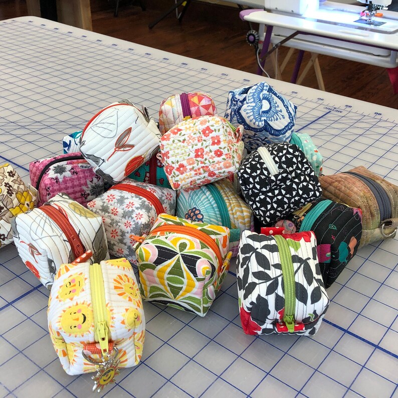 May include: A collection of 14 small, colorful fabric cubes with zipper closures. The cubes are made of various patterned fabrics, including floral, geometric, and abstract designs.