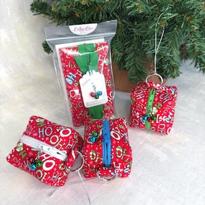 May include: Three small, square, fabric zipper pouches featuring a red background with a repeating pattern of green and white 'HO' text and candy cane motifs. Each pouch has a colorful zipper and a small metal jingle bell charm attached to the pull.
