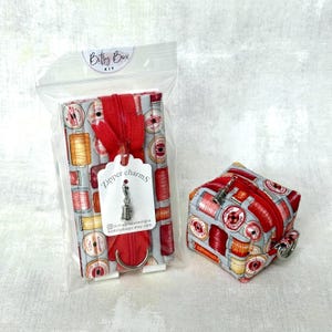 May include: A rectangular fabric pouch and a small square fabric pouch, both featuring a print of red and yellow thread spools. The rectangular pouch is inside a clear plastic bag with a tag labeled Zipper charms and a metal spool charm attached.