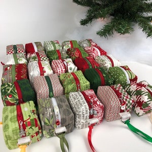 May include: A collection of 24 small, handmade zippered pouches in various Christmas-themed fabrics. The pouches are made with red, green, and white fabrics with patterns of holly, snowflakes, and Christmas words. Each pouch has a zipper closure and a small loop for hanging.