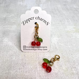 May include: Two gold zipper charms with red and green cherry designs. The charms are attached to a gold chain and have a gold lobster clasp. The charms are displayed on a white card with the text "Zipper charms" and the social media handle "@ticklegrassdesigns bodobobags.etsy.com".