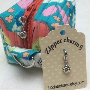May include: A small, quilted zipper pouch with a floral print in shades of pink, blue, and green. The pouch has a silver zipper pull with a flower charm attached. A tag is attached to the pouch that reads "Zipper charms" and "bodobobags.etsy.com".