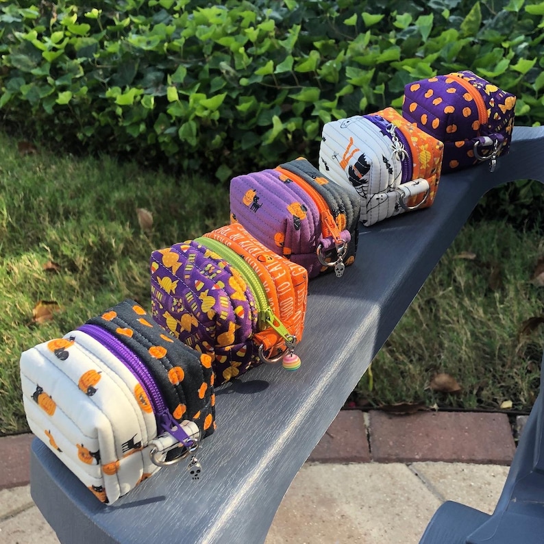 May include: Five small Halloween-themed zipper pouches with orange, purple, black, and white fabric. The pouches are lined up on a gray surface and have small charms attached to the zippers.
