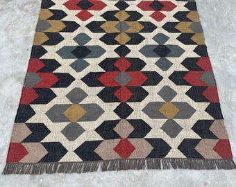 May include: A rectangular area rug featuring a repeating geometric pattern of diamonds and hexagons in shades of red, mustard yellow, charcoal gray, and beige on an off-white background, finished with a short gray fringe along the bottom edge.