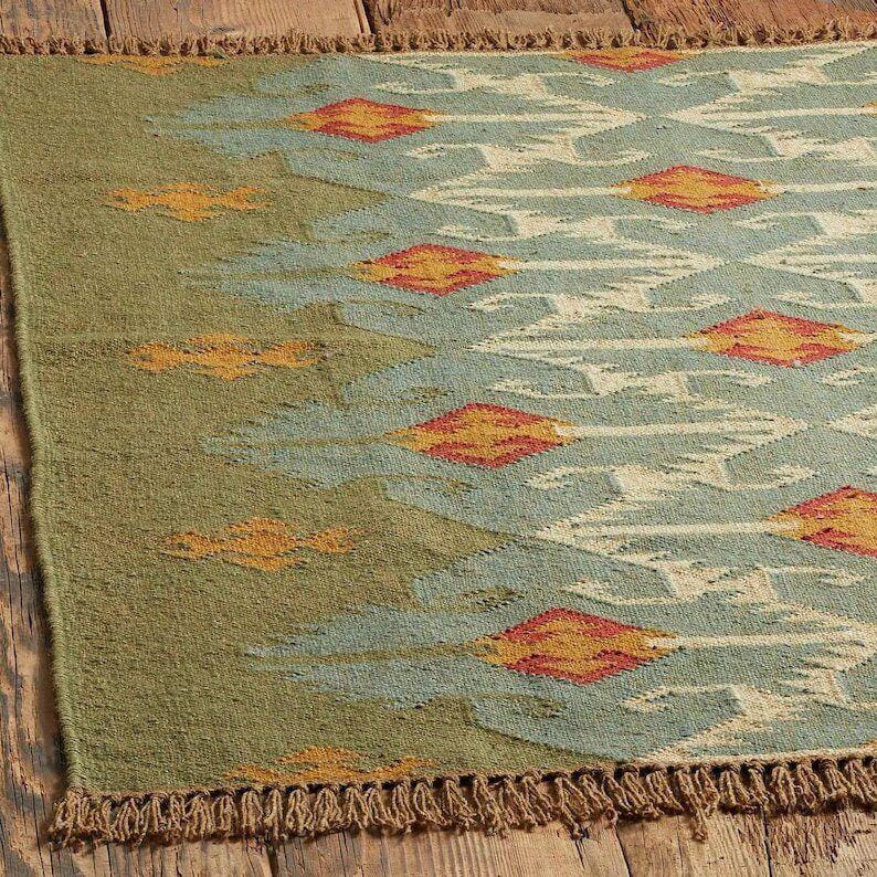 May include: A rectangular woven rug featuring a muted olive green base color with a repeating pattern of light blue diamond shapes. Each diamond contains a smaller orange and red geometric center. The rug has a textured fringe along the edges.