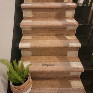 Puede incluir: Una alfombra de escalera de yute tejida con una textura bronceada natural y bloques rectangulares blancos alternos a lo largo de los bordes. La alfombra cubre varios escalones y el texto TejRugsExport es visible en uno de los peldaños.