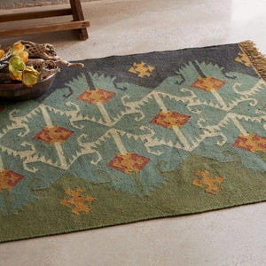 May include: A rectangular area rug featuring a repeating geometric pattern in shades of sage green, dark charcoal, and muted orange. The design includes stylized diamond shapes and small cross motifs, finished with a short fringe on one end.