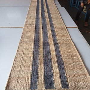 Peut inclure: Un long tapis de couloir rectangulaire tissé avec une base en fibres naturelles de couleur beige et trois bandes verticales gris anthracite foncé au centre. La texture est grossière et tressée, avec le texte Tej Rugs Export dans le coin.