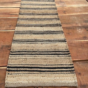 May include: A rectangular woven runner rug featuring a natural tan fiber texture with repeating horizontal black stripes throughout the length. The rug has a visible coarse weave pattern and includes the text TejRugsExport at the bottom.