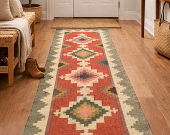 May include: A long rectangular runner rug featuring a geometric pattern with a central red field, cream-colored borders, and accents in olive green, pink, and dark gray. The rug displays a repeating diamond motif and the text Tej Rugs Export.