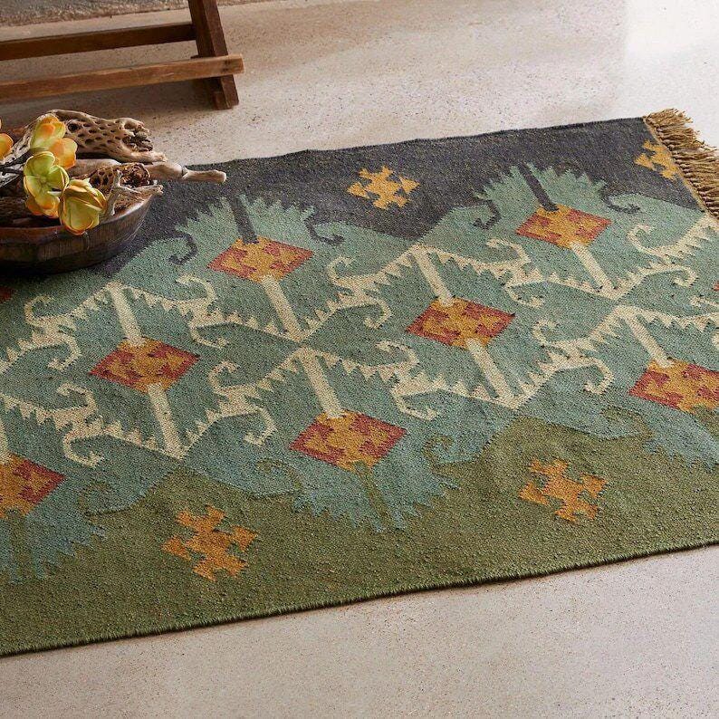 May include: A rectangular area rug featuring a repeating geometric pattern in shades of sage green, dark charcoal, and muted orange. The design includes stylized diamond shapes and small cross motifs, finished with a short fringe on one end.