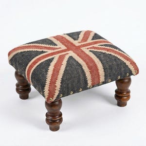 May include: A rectangular footstool featuring a woven Union Jack pattern in red, white, and dark grey. The top is secured with metallic nailhead trim along the edges and is supported by four dark brown, turned wooden legs with a polished finish.
