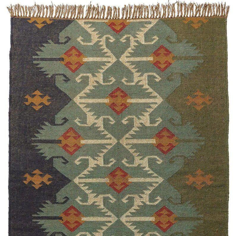 May include: A rectangular woven rug featuring a central vertical band of teal with geometric patterns in cream, red, and orange. The left side is dark charcoal and the right side is olive green, both with small orange motifs. The top edge has fringe.