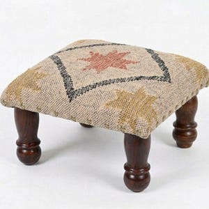 May include: A low footstool with a square, textured beige top featuring a central diamond pattern in black with a red star motif and additional gold star accents. The stool is supported by four dark brown, turned wooden legs with a rounded base.