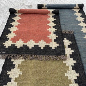 Puede incluir: Tres alfombras rectangulares tejidas con bordes de flecos, que presentan un bloque de color sólido central en terracota, verde oliva y azul pizarra, rodeado por un borde negro grueso con un patrón geométrico de cruces en color crema.