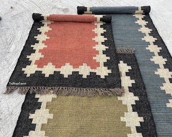 May include: Three rectangular woven rugs with fringed edges, featuring a central solid color block in terracotta, olive green, and slate blue, surrounded by a thick black border with a repeating cream-colored geometric cross pattern.