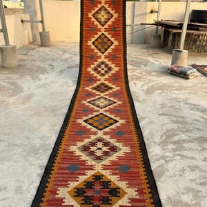 May include: A long, narrow runner rug featuring a repeating geometric diamond pattern in shades of rust orange, mustard yellow, black, and cream. The rug has a dark border and includes the text Tej Rugs Export printed near the bottom edge.