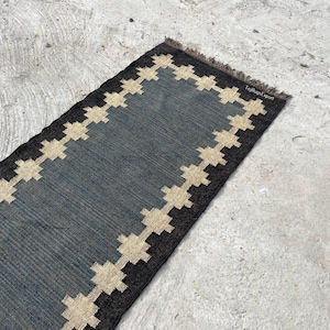 Può includere: Un tappeto rettangolare a passatoia con un centro blu scuro dalla trama testurizzata. Il tappeto è bordato da un motivo geometrico ripetitivo a croce in color beige chiaro, rifinito con frange marrone scuro sul bordo corto.
