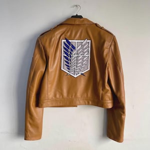 May include: A tan leather jacket featuring a large embroidered emblem on the back. The emblem displays two stylised wings, one in blue and one in white, set against a grey shield background. The jacket has a classic collar and long sleeves.