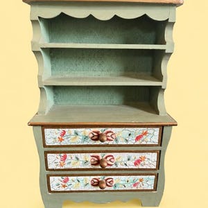 May include: A sage green wooden hutch with a natural wood top, featuring two open shelves and three drawers. Each drawer front displays a white background with a crackle finish, decorated with painted red and orange floral motifs and round knobs.