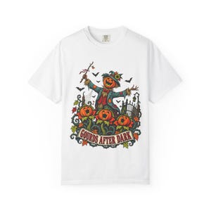Peut inclure: Un t-shirt blanc à manches courtes orné d'un motif représentant un épouvantail à tête de citrouille en manteau bleu, entouré de trois citrouilles chantantes, de chauves-souris et de feuilles d'automne, avec le texte GOURDS AFTER DARK.