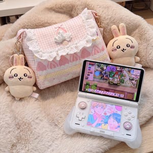 Puede incluir: Un bolso bandolera de cuadros vichy rosa y blanco con ribete de encaje y motivos de fresas, dos colgantes de conejo de peluche y una consola de juegos portátil blanca que muestra una ilustración anime y una interfaz de juego en las pantallas.