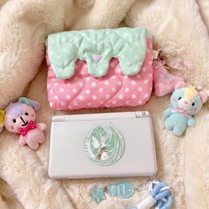 Puede incluir: Un estuche acolchado rosa con lunares blancos y una solapa festoneada verde menta con un pequeño corazón, acompañado de una consola de juegos portátil blanca con una pegatina verde circular, dos colgantes de peluche y un cordón azul con etiqueta.