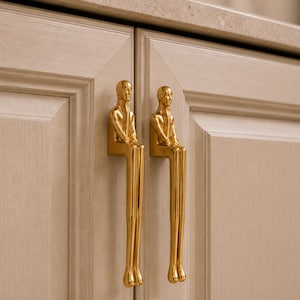 May include: Two gold-colored cabinet pulls shaped like stylized human figures sitting with their legs dangling downward. The figures have smooth, reflective surfaces and are mounted vertically on light-colored wooden cabinet doors with a paneled design.