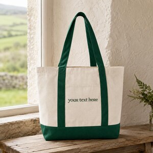 May include: A cream-colored canvas tote bag featuring dark green trim at the base and two matching dark green shoulder straps. The front of the bag displays the text "your text here" in a dark green, simple, sans-serif font centered on the panel.