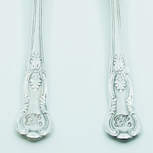 May include: Two silver spoons with ornate handles and a reflective surface. The handles feature a floral design and a monogram. The spoons are displayed on a white background, showcasing their elegant design and craftsmanship.