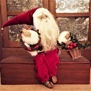 May include: A Santa Claus figurine with a long white beard, red velvet hat and pants, and a black and gold jacket. He holds a small wreath and a basket of red apples. The figurine is seated on a wooden surface.
