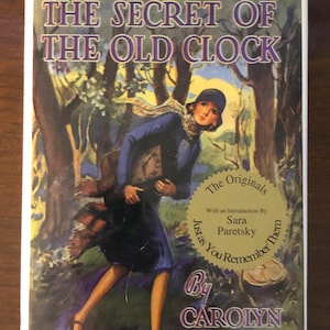 May include: A vintage book cover featuring a woman in a blue coat and hat, holding a book, walking through a forest. The title of the book is "The Secret of the Old Clock" by Carolyn Keene. The cover also includes the text "Nancy Drew Mystery Stories" and "The Originals, Just as You Remember Them".