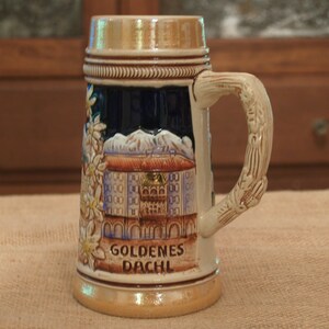 May include: A ceramic beer stein with a brown, gold, and blue design. The stein features a depiction of a building with a golden roof and the text "GOLDENES DACHL".