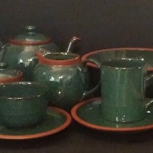 May include: A green tea set with orange trim. The set includes a teapot, sugar bowl, creamer, cup and saucer, and a serving bowl. The tea set is arranged on a dark surface.