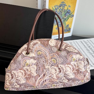Puede incluir: Un bolso de mano semicircular con un intrincado patrón floral y abstracto en tonos marrón, crema y morado suave. El bolso tiene dos asas finas y redondeadas de color marrón oscuro unidas con ojales metálicos dorados y cierre de cremallera.