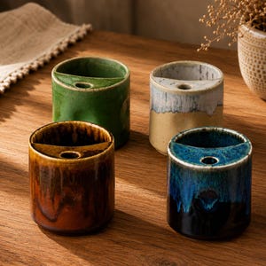 Pode incluir: Quatro porta-escovas de dentes cilíndricos de cerâmica com um divisor interno semicircular e uma pequena abertura redonda. Os suportes apresentam esmaltes reativos distintos em verde, bege e branco, castanho e âmbar, e azul e preto.