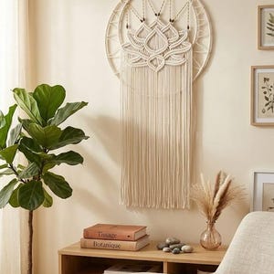 Può includere: Una decorazione da parete circolare in macramè con un motivo a fiore di loto color bianco sporco realizzato in corda annodata. L'oggetto include un grande anello esterno, perline di legno decorative e lunghe frange dritte pendenti.