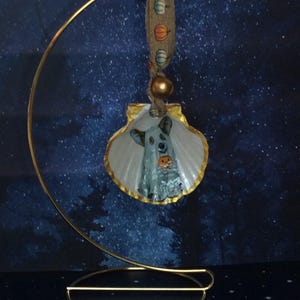 May include: A white seashell ornament with a gold-painted edge, featuring a painted ghost dog holding a small orange pumpkin. The shell hangs from a tan ribbon patterned with tiny pumpkins, suspended on a curved gold-toned metal display stand.