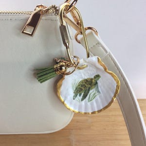 May include: A white handbag featuring a gold-toned metal carabiner clip attached to the strap. A white seashell charm with a painted green sea turtle and gold-painted edges hangs from the clip, accompanied by a small sage green tassel.