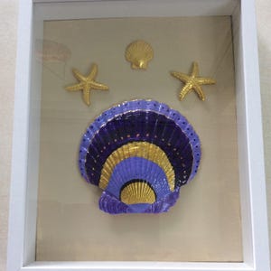 May include: A white shadow box frame containing a large, central scallop shell painted in purple, black, and gold. Above the large shell are two gold starfish and one smaller gold scallop shell, all arranged on a light beige backing board.